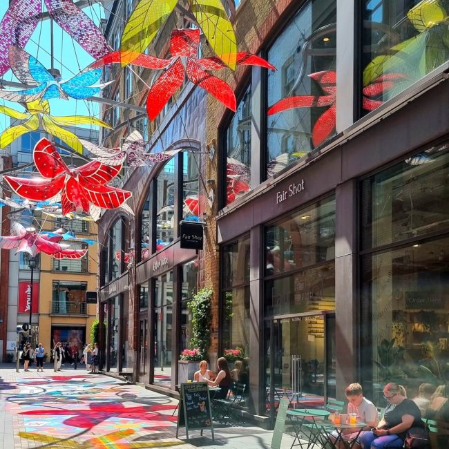 St Martins Courtyard with large decorative flowers suspended between buildings while people sit at cafe tables underneath